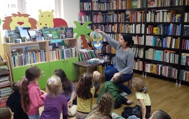 Dzieci siedzą na dywanie na przeciwko bibliotekarki i patrzą jak bibliotekarka pokazuje im książeczkę pluszową.