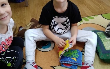 Chłopiec o blond włosach siedzi na dywanie i przegląda pluszową książeczkę