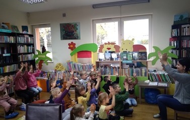 Dzieci siedzą na dywanie i mają ręce w g&oacute;rze naśladując to co pokazuje Pani bibliotekarka, kt&oacute;ra siedzi na przeciwko nich.