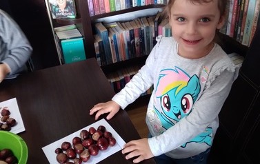 Dziewczynka stoi przy stoliku, na stole leży kartka obłożona kasztanami według wzoru.