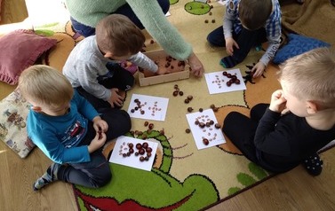 Chłopcy siedzą na dywanie i układają na kartkach kasztany według wzoru. W tle widać rękę opiekuna.
