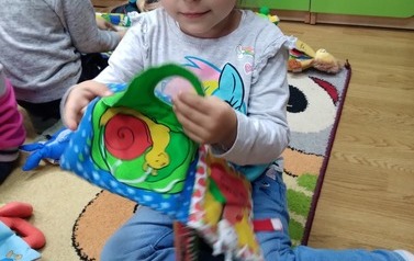 Dziewczynka o blond włosach siedzi w bibliotece na dywanie i ogląda pluszową książeczkę.