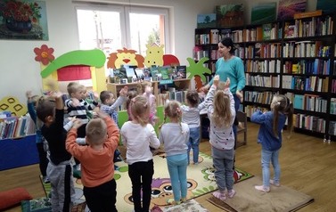 Dzieci stoją w rozsypce twarzą do bibliotekarki z podniesionymi rękami do g&oacute;ry naśladując to co Pani pokazuje.