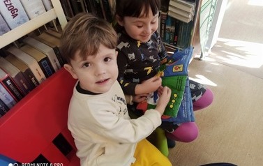 Dziewczynka i chłopiec siedzą na krzesełkach i  razem przeglądają pluszową książeczkę.