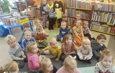 Grupa dzieci siedzi na dywanie skierowana twarzą do aparatu, a po środku siedzi bibliotekarka i obejmuje dwie dziewczynki.