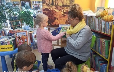 Pani bibliotekarka trzyma pluszową książeczkę i stoi przy niej dzieczynka w r&oacute;żowym sweterku i pokazuje coś rączką w książeczce.  Widać kilkoro dzieci.