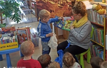 Pani bibliotekarka trzyma pluszową książeczkę i stoi przy niej chłopiec, kt&oacute;ry wyjmuje z książeczki  rybkę aby włożyć ją do siateczki, kt&oacute;rą trzyma w prawej dloni. Widać kilkoro dzieci.