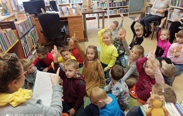 Dzieci siedzą na dywanie, niekt&oacute;re z nich zgłaszają się aby odpowiedzieć na zagadkę powiedzianą przez Panią bilbiotekarkę.Bibliotekarka siedzi na krzesełku po środku i czyta z kartki zagadki.