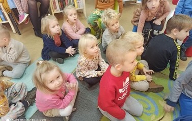 Dzieci siedzą na dywanie i słuchają bajkę czytaną przez bibliotekarkę.