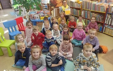 Grupa dzieci siedząca na dywanie, z tyłu Pani bibliotekarka z książką w rękach - pozują do zdjęcia grupowego.