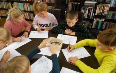 Dzieci siedzą przy stole w bibliotece i każdy ma swoją jesienną kartę pracy. Na stole leżą kredki.