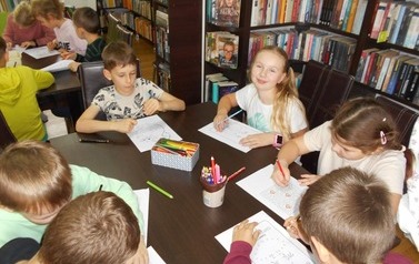 Dzieci siedzą przy stole w bibliotece i każdy ma swoją jesienną kartę pracy. Na stole leżą kredki.