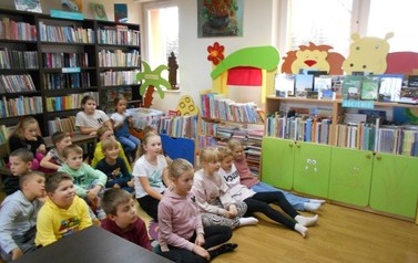 Dzieci siedzą w grupie na dywanie i patrzą na bibliotekarkę.