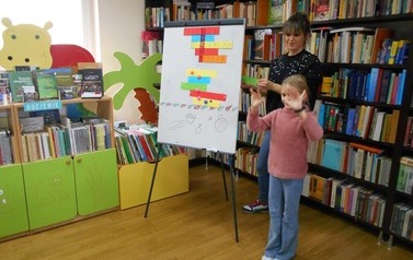 Przy tablicy magnetycznej stoi bibliotekarka z dziewczynką, kt&oacute;ra pokazuje coś rękoma do dzieci.