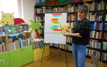 Bibliotekarka stoi przy magnetycznej tablicy na kt&oacute;rej są kolorowe paski. W ręku trzyma książkę bajkową.