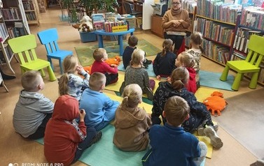 Dzieci siedzą na karimatach i patrzą na Panią bibliotekarkę i słuchają co m&oacute;wi.