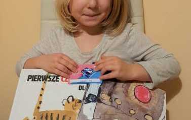 Dziewczynka o blond włosach siedzi na fotelu trzymając swoją nową wyprawkę czytelniczą