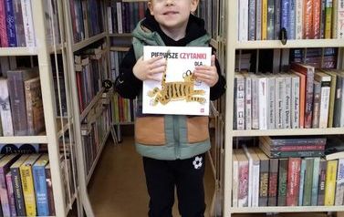 Chłopiec o blond włosach stoi ze swoją nową książką w bibliotece w Zblewie.