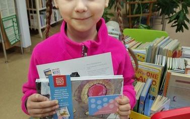 Dziewczynka w r&oacute;żowej bluzce stoi w bibliotece w Zblewie ze swoją wyprawką czytelniczą.