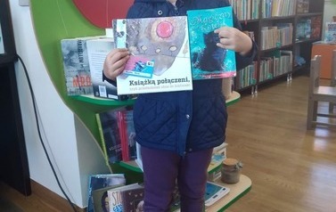Dziewczynka o blond włosah stoi ze swoją wyprawką czytelniczą i swoją książką wypożyczoną w bibliotece w Pinczynie