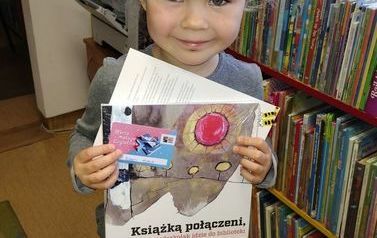 Dziewczynka o blond włosach stoi ze swoją wyprawką czytelniczą.