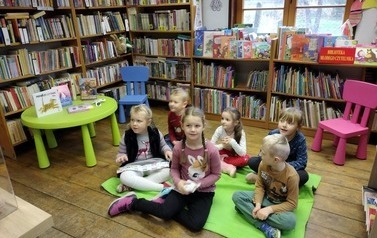 Dzieci siedzą na kocu w bibliotece w Borzechowie i słuchają bibliotekarki.