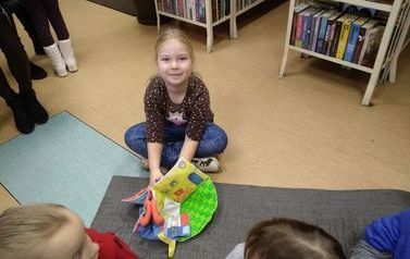 Dziewczynka o blond włosach siedzi na podłodze i bawi się pluszową książeczką. W tle widać inne dzieci.