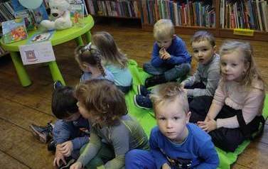 Dzieci siedzą na dywanie, obok widać stolik okrągły z misiem i książeczkami i trzy balony.