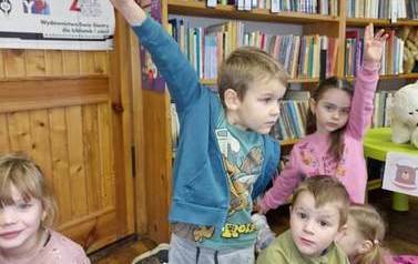 Dzieci siedzą na kocykach w bibliotece, chłopiec i dziewczynka zgłaszają się podnąsząc rękę.