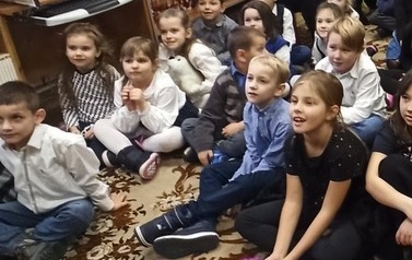 Grupa dzieci wraz z opiekunami siedzi na dywanie i ogląda teatrzyk.