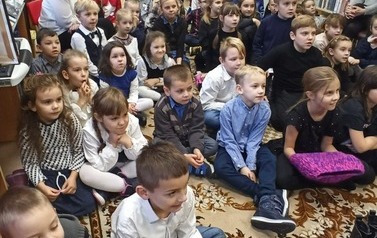 Duża grupa dzieci wraz z opiekunami siedzi na dywanie i ogląda teatrzyk.