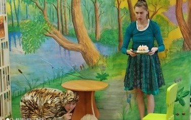 Dziewczyna trzyma w ręku tort, a pod stolikiem chowie się tygrysek. W tle las z potokiem.