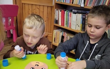 Dw&oacute;ch chłopc&oacute;w przy okrągłym stoliku wykleja papierowego misia.