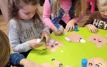 Dzieci stoją przy okrągłym zielonym stoliku i wyklejają papierowego misia.