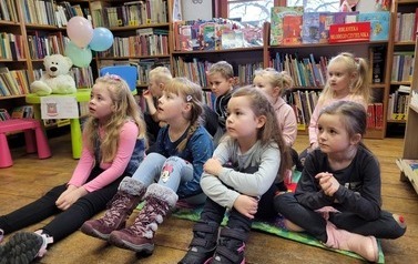 Grupa dzieci siedzi na kocykach i słucha bibliotekarza.