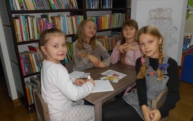 Dziewczynki siedzą przy stoliku i wyklejają puzzle misia.Za nimi na tablicy jest narysowany duży miś.