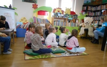 Dzieci siedzą na dywanie i słuchają bajki czytanej przez bibliotekarkę. Z prawej strony na krześle siedzi duży miś.