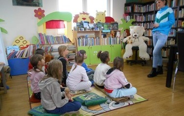 Dzieci siedzą na dywanie i patrzą na bibliotekarkę, kt&oacute;ra opowiada o Dniu Misia. Z boku widać dużego misia siedzącego na krześle.