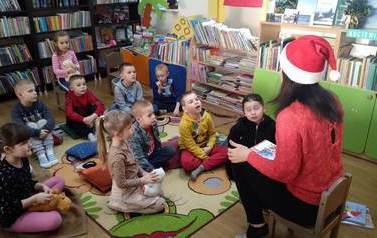 Dzieci siedzą na dywanie i słuchają tego co m&oacute;wi bibliotekarka, kt&oacute;ra ubrana jest w czapkę świętego Mikołaja.