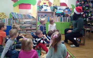 Dzieci siedzą na dywanie,a bibliotekarka  w czapce Mikołaja czyta im bajkę.