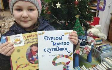 Dziewczynka w czapce w paski pokazuje sw&oacute;j dyplom Małego Czytelnika, książkę i naklejkę.