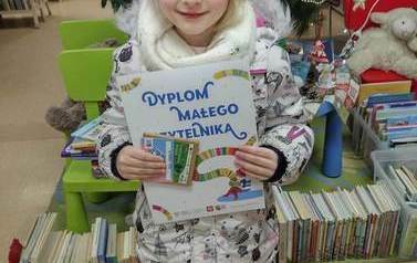 Dziewczynka w r&oacute;żowej czapce pokazuje sw&oacute;j dyplom Małego Czytelnika, w rączce też trzyma kredki i kartę magnetyczną. 