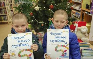 Dw&oacute;ch chłopc&oacute;w pokazuje swoje dyplomy Małego Czytelnika. W tle widać choinkę.