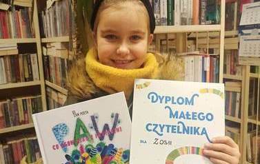 Dziewczynka o blond włosach pokazuje sw&oacute;j Dyplom Małego Czytelnika i wypożyczoną książeczkę