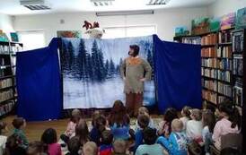 Na tle zaśnieżonego lasu stoi aktor i patrzy w g&oacute;rę na głowę renifera i rozmawiają. Publiczność dziecięca patrzy.