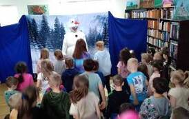 Na tle śnieżnego lasu stoi bałwanek Olaf, a dzieci na Niego patrzą i go naśladują stojąć i ruszając się tak jak On.