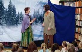 Na tle leśnego, zimowego krajobrazu stoi aktorka ubrana w zieloną sukienkę z fioletową peleryną i rozmawia z aktorem ubranym w beżową tunikę i brązowe spodnie i z czapką na uszach.Widać publiczność dziecięcą.