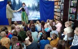 Na tle zimowego, leśnego krajobrazu stoją aktor i aktorka teatru trzymając się za prawą dłoń. Publicznośc dziecięca patrzy na nich.