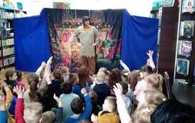 Z przodu stoi aktor z ręcami załozonymi na biodrach i patrzy na dziecięcą publiczność, z ktorych niekt&oacute;re dzieci mają podniesione ręce aby coś powiedzieć.