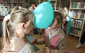 Dwie dziewczynki stoją w parze twarzami do siebie i trzymając się za ręce trzymają głowami zielony balon.